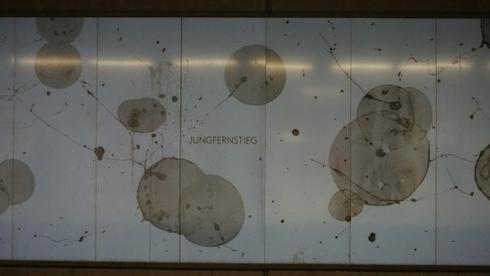 Circular patterns and splatters resembling abstract art are displayed on a light background with the word 'JUNGERNSTIEG' in the center. The design features various shades of gray and brown, creating a modern and minimalist aesthetic.