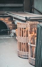 Two large wicker baskets filled with freshly baked baguettes are placed on a tiled floor near a brick oven. The oven has a metallic surface and a partly open door, suggesting recent use.