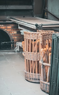 Two large wicker baskets filled with freshly baked baguettes are placed on a tiled floor near a brick oven. The oven has a metallic surface and a partly open door, suggesting recent use.