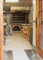 A rustic bakery interior featuring a large brick oven, with several shelves and baking tools including long wooden paddles hanging from the ceiling. Baskets are filled with baguettes or similar bread, and some wrapped items are on the tiled floor. The space has a well-used, functional appearance.