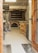 A rustic bakery interior featuring a large brick oven, with several shelves and baking tools including long wooden paddles hanging from the ceiling. Baskets are filled with baguettes or similar bread, and some wrapped items are on the tiled floor. The space has a well-used, functional appearance.