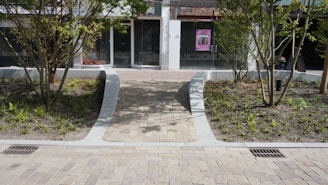 A stone pathway leads to a glass door entrance of a building, with planted areas featuring young trees and small shrubs on both sides. The facade has a modern appearance with a large glass window and a visible poster on the door.