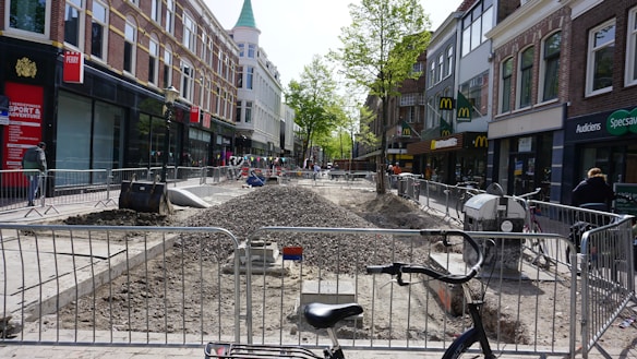 A street undergoing construction with barriers separating a central area filled with gravel. Several shops are visible on both sides, including a fast-food restaurant. There are trees in the background and a bicycle is parked in the foreground.