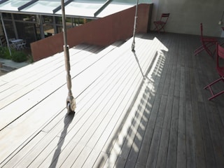 A wooden deck area is partially in sunlight and partially shaded. Two metal supports are casting long shadows across the deck. In the background, there are glass windows and a small outdoor seating area with white furniture. The deck area features red folding chairs against a light green wall.
