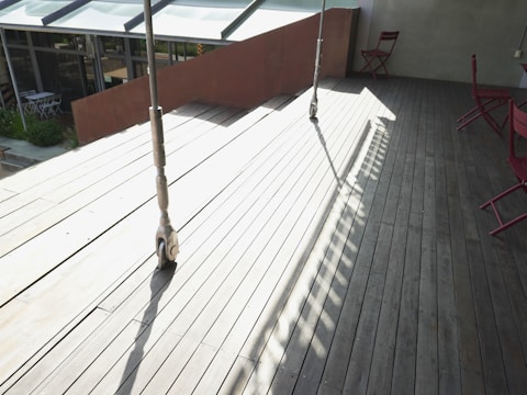 A wooden deck area is partially in sunlight and partially shaded. Two metal supports are casting long shadows across the deck. In the background, there are glass windows and a small outdoor seating area with white furniture. The deck area features red folding chairs against a light green wall.