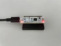 A small electronic circuit board with various components is placed on a black plastic base. It is connected to a USB cable, with two red indicator lights illuminated on its surface.