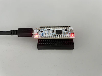 A small electronic circuit board with various components is placed on a black plastic base. It is connected to a USB cable, with two red indicator lights illuminated on its surface.
