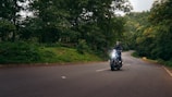 A motorcyclist rides along a winding road surrounded by lush green trees and dense forest. The road curves ahead into the distance, and the rider is wearing a helmet and protective gear. Soft daylight filters through the canopy, creating a serene and adventurous setting.