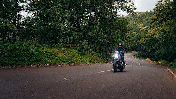 A motorcyclist rides along a winding road surrounded by lush green trees and dense forest. The road curves ahead into the distance, and the rider is wearing a helmet and protective gear. Soft daylight filters through the canopy, creating a serene and adventurous setting.