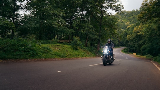 A motorcyclist rides along a winding road surrounded by lush green trees and dense forest. The road curves ahead into the distance, and the rider is wearing a helmet and protective gear. Soft daylight filters through the canopy, creating a serene and adventurous setting.