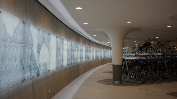 A modern indoor bicycle parking station with sleek architectural design. Bicycles are stacked neatly on multi-level racks to the right, while the left side features a long, illuminated panel displaying abstract or stylized artwork. The ceiling is smooth and features recessed lighting, creating a clean and organized environment.