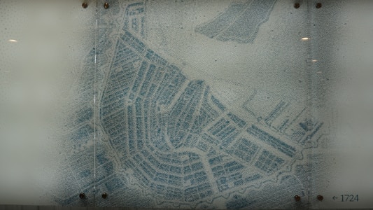A detailed map with intricate blue lines displays a network of streets and canals, resembling the layout of a historical city or district. The map is printed on a slightly translucent surface with its corners fastened by screws. The number '1724' is visible in the bottom right, potentially indicating a date or map reference.