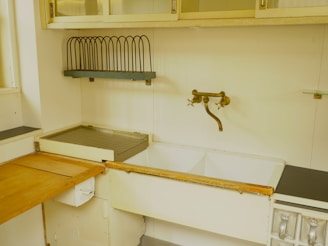 A vintage kitchen sink setup featuring a double basin with a rustic bronze faucet mounted on the wall. Above the sink, there is a metal dish rack for drying dishes and cabinets with glass doors. The countertop is a mix of wood and metal, displaying signs of wear. The color palette is muted, giving it a retro vibe.