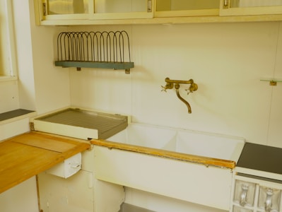 A vintage kitchen sink setup featuring a double basin with a rustic bronze faucet mounted on the wall. Above the sink, there is a metal dish rack for drying dishes and cabinets with glass doors. The countertop is a mix of wood and metal, displaying signs of wear. The color palette is muted, giving it a retro vibe.