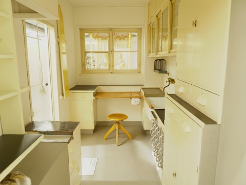 A vintage-looking kitchen space with cream-colored cabinetry and counters alongside a small wooden stool. The room features a window with decorative curtains, a sink area with a brass faucet, and orderly arranged cabinets and drawers. The color palette is predominantly soft yellow and cream, creating a nostalgic atmosphere.