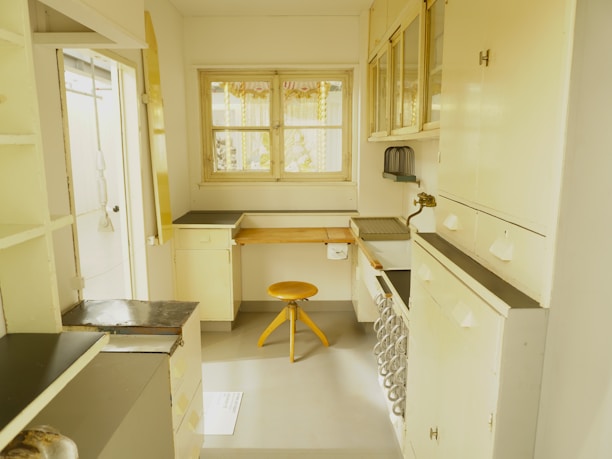 A vintage-looking kitchen space with cream-colored cabinetry and counters alongside a small wooden stool. The room features a window with decorative curtains, a sink area with a brass faucet, and orderly arranged cabinets and drawers. The color palette is predominantly soft yellow and cream, creating a nostalgic atmosphere.
