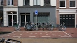 Several bicycles are parked in an organized manner on a brick-paved street in front of a row of urban buildings. The buildings have a mix of white, gray, and dark green colors with large windows and doors. A parking sign is visible above the bikes, indicating a designated parking area. The street is clean and appears to be in a residential or mixed-use neighborhood.
