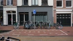 Several bicycles are parked in an organized manner on a brick-paved street in front of a row of urban buildings. The buildings have a mix of white, gray, and dark green colors with large windows and doors. A parking sign is visible above the bikes, indicating a designated parking area. The street is clean and appears to be in a residential or mixed-use neighborhood.