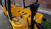 A yellow bike-like vehicle with multiple seats and seat belts is parked next to a brick wall. The vehicle features a steering column with various controls and is designed for group transportation. The ground is paved, and there is some greenery visible in the background.