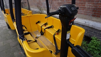 A yellow bike-like vehicle with multiple seats and seat belts is parked next to a brick wall. The vehicle features a steering column with various controls and is designed for group transportation. The ground is paved, and there is some greenery visible in the background.