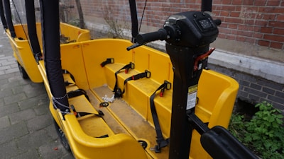 A yellow bike-like vehicle with multiple seats and seat belts is parked next to a brick wall. The vehicle features a steering column with various controls and is designed for group transportation. The ground is paved, and there is some greenery visible in the background.