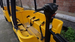 A yellow bike-like vehicle with multiple seats and seat belts is parked next to a brick wall. The vehicle features a steering column with various controls and is designed for group transportation. The ground is paved, and there is some greenery visible in the background.