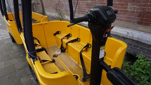 A yellow bike-like vehicle with multiple seats and seat belts is parked next to a brick wall. The vehicle features a steering column with various controls and is designed for group transportation. The ground is paved, and there is some greenery visible in the background.