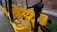 A yellow bike-like vehicle with multiple seats and seat belts is parked next to a brick wall. The vehicle features a steering column with various controls and is designed for group transportation. The ground is paved, and there is some greenery visible in the background.