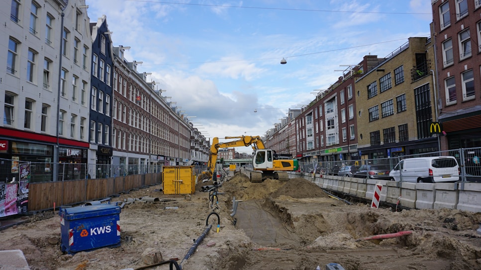 A street undergoing construction, with a yellow excavator in the center lifting dirt. Surrounding the construction site are tall buildings on both sides of the street, with various shops and parked vehicles. There is a blue container labeled 'KWS' on the left and construction materials scattered throughout the area. The sky is partly cloudy.