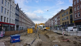 A street undergoing construction, with a yellow excavator in the center lifting dirt. Surrounding the construction site are tall buildings on both sides of the street, with various shops and parked vehicles. There is a blue container labeled 'KWS' on the left and construction materials scattered throughout the area. The sky is partly cloudy.
