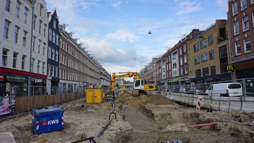 A street undergoing construction, with a yellow excavator in the center lifting dirt. Surrounding the construction site are tall buildings on both sides of the street, with various shops and parked vehicles. There is a blue container labeled 'KWS' on the left and construction materials scattered throughout the area. The sky is partly cloudy.