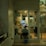 A security guard is seated inside a small, glass-enclosed security booth. The booth is filled with various monitoring and communication equipment, including multiple surveillance screens, a fan, and documents pinned on the walls. The setting appears to be in a dimly lit area, giving off an evening or night-time ambiance.