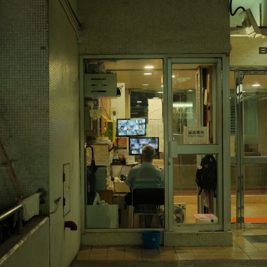 A security guard is seated inside a small, glass-enclosed security booth. The booth is filled with various monitoring and communication equipment, including multiple surveillance screens, a fan, and documents pinned on the walls. The setting appears to be in a dimly lit area, giving off an evening or night-time ambiance.