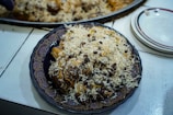 A plate of rice mixed with what appears to be raisins and some saffron strands, topped over a piece of meat on a decorative plate. The dish is placed on a white countertop next to a stack of plates.