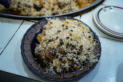 A plate of rice mixed with what appears to be raisins and some saffron strands, topped over a piece of meat on a decorative plate. The dish is placed on a white countertop next to a stack of plates.