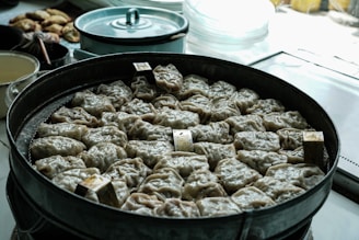 A large metal steamer filled with neatly arranged dumplings, each with a crinkled appearance, indicating they are ready to be cooked or recently cooked. The background displays various kitchen items including a light blue pot, suggesting a culinary setting.