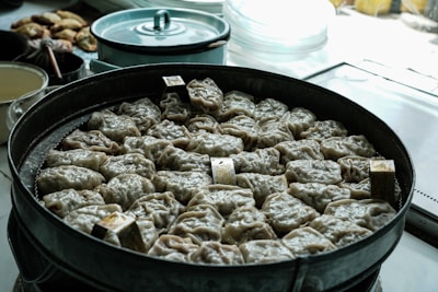 A large metal steamer filled with neatly arranged dumplings, each with a crinkled appearance, indicating they are ready to be cooked or recently cooked. The background displays various kitchen items including a light blue pot, suggesting a culinary setting.