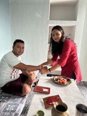 A woman in a red kurta is tying a rakhi on a man's wrist. They are sitting at a table with a plate of sweets and a gift envelope. The setting suggests a celebration or traditional ceremony.