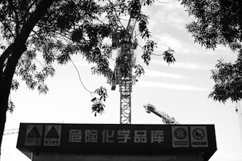 A black and white image featuring a construction crane towering in the background, partially obscured by tree branches. The foreground contains a sign with Chinese characters and warning symbols.