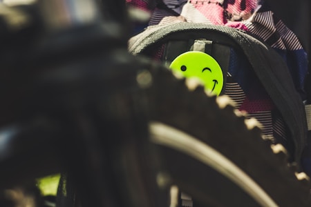 A bright green winking smiley face badge is attached to a backpack. The foreground features part of a bicycle wheel, which is slightly blurred. The backpack has a colorful, patterned fabric, creating a contrast with the neon green badge.