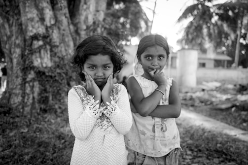 Two young children stand side by side with thoughtful expressions and their hands touching their faces. They are positioned in front of large tree trunks, with blurry foliage and structures visible in the background. The setting appears outdoors and natural.