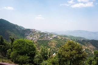 A picturesque landscape featuring a village spread across a lush green hillside, with mountains in the background. The village houses are interspersed with trees and vegetation, creating a harmonious blend of nature and human settlement. The sky is bright and clear, adding to the serene atmosphere.