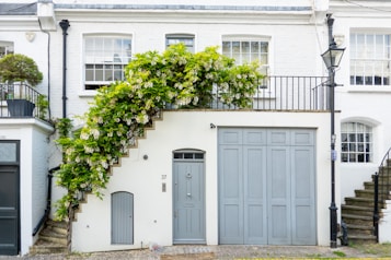 A charming white brick townhouse features a spiral staircase adorned with lush greenery and flowers. The property includes large windows, a blue-gray door, and a matching garage door. A vintage lamp post stands on the stone pathway in front, adding to the quaint atmosphere.