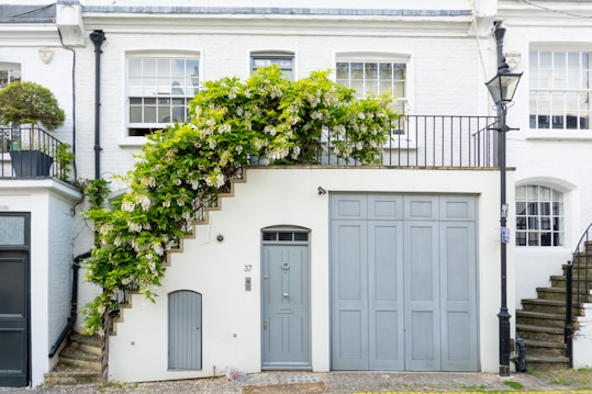 A charming white brick townhouse features a spiral staircase adorned with lush greenery and flowers. The property includes large windows, a blue-gray door, and a matching garage door. A vintage lamp post stands on the stone pathway in front, adding to the quaint atmosphere.