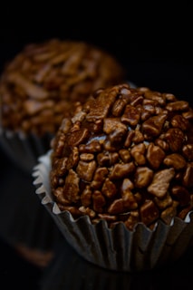 Two chocolate truffle candies with a textured coating of chocolate pieces are presented in silver cupcake liners against a dark background.
