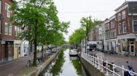 A canal flanked by buildings and lined with green trees. Several bicycles are parked along the sidewalk, and a small boat is moored on the water. The scene features a blend of historic architecture and nature, creating a tranquil urban atmosphere.