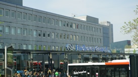 A busy transportation hub features a large, modern building with numerous windows. A sign in the foreground reads 'Eindhoven Centraal'. There are people gathered near the entrance and a bus visible, indicating a public transit area. The scene is bright with some greenery visible on the right side.