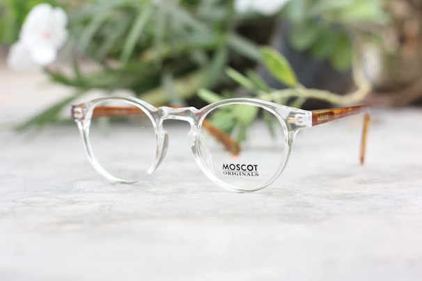 A pair of clear eyeglasses with brown temples rests on a light-colored surface, with blurred green foliage in the background. The glasses have a distinctive round frame design typical of classic optical styles.