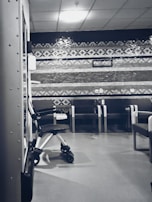 A clinical waiting area with a row of empty chairs against a wall decorated with geometric patterns. A wheelchair is parked to one side, and the lighting is soft and subdued. The setting seems orderly and quiet with a sterile ambiance.