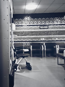 A clinical waiting area with a row of empty chairs against a wall decorated with geometric patterns. A wheelchair is parked to one side, and the lighting is soft and subdued. The setting seems orderly and quiet with a sterile ambiance.